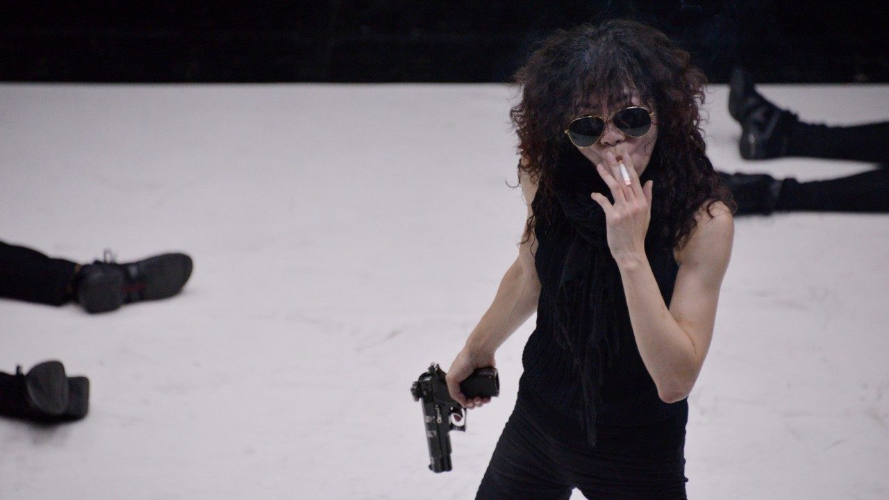 An Asian woman with long wavy hair wears all black and aviator sunglasses. She takes a drag on a cigarette and holds a gun in her hand. Behind her are two pairs of legs laying on the floor.