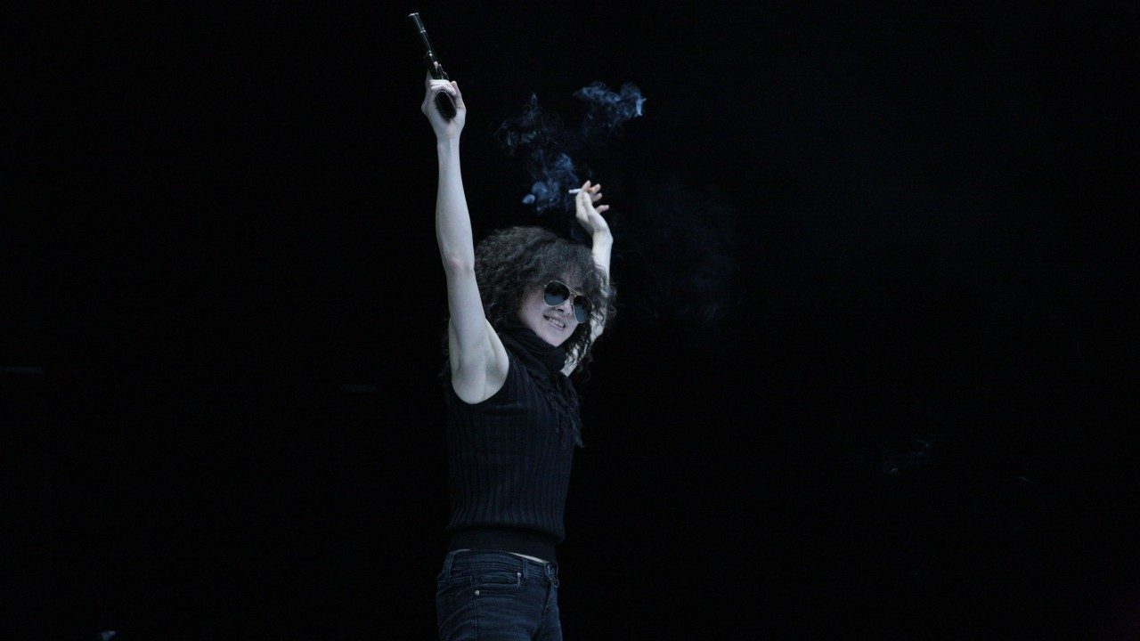  An Asian woman wearing a black sleeveless turtleneck and dark jeans stands on stage in front of a black background, both arms lifted above her head. In one hand she holds a gun, in the other, a cigarette. She smiles widely below her aviator sunglasses.
