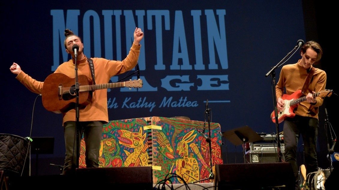  Two young white men perform on stage during a Mountain Stage concert. The man on the left wears a camel colored sweater with an acoustic guitar hanging across his body by its strap. He sings passionately with his eyes closed, arms outstretched. At right, a white man with medium length hair wears a camel colored shirt and plays a red electric guitar.