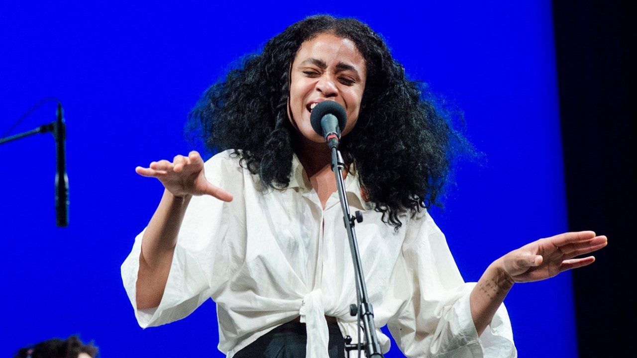  A Black woman with long, wavy natural hair parted down the middle sings into a microphone. She wears a white shirt and hold both hands out in front of her as she hits her note, a blue background behind her.