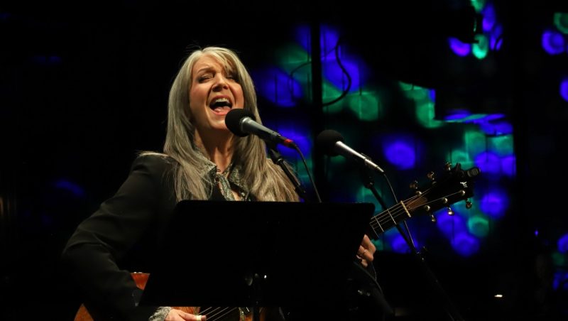  Host Kathy Mattea sings into a microphone on stage during a Mountain Stage performance. Mattea is a white woman with long grey hair. She wears a black blazer and plays an acoustic guitar, which is just visible behind a music stand. The background is ethereal-looking blue and grey lights.