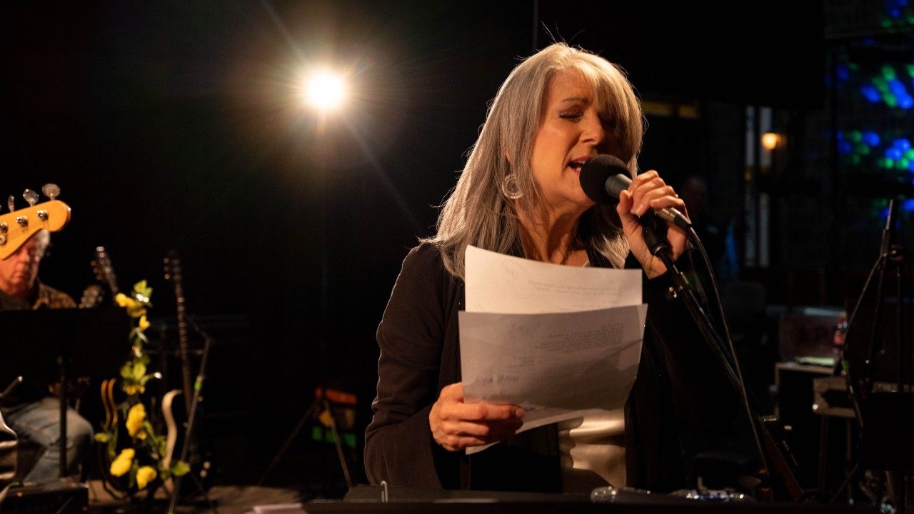  Host Kathy Mattea sings into a microphone and holds two sheets of paper. Musicians perform nearby in the background. Mattea is a white woman with medium length grey hair. She wears a black shirt and large silver earrings.