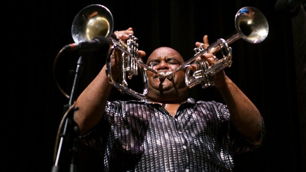  A man bald man from Dirty Dozen Brass Band plays two trombones at once, beads of sweat visible on his head from performing. He wears a patterned grey shirt in front of a black background.