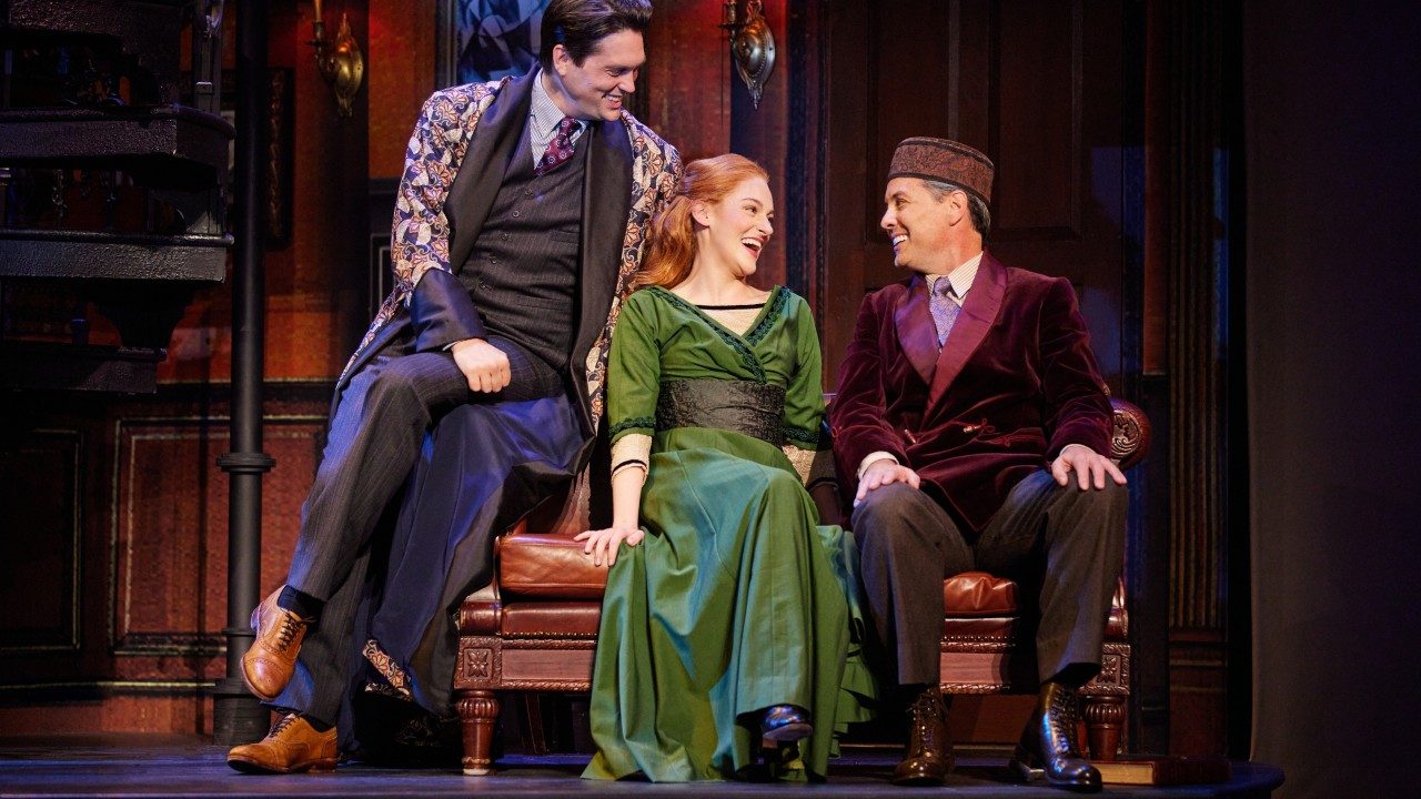  Jonathan Grunert as Professor Henry Higgins, Madeline Powell as Eliza Doolittle, and John Adkinson as Colonel Pickering in the national tour of "My Fair Lady." Photo by Jeremy Daniel. Two white men in early 1900s smoking jackets flank a redheaded white woman in a green gown. They smile towards her and she laughs, all sitting on a cognac leather loveseat.