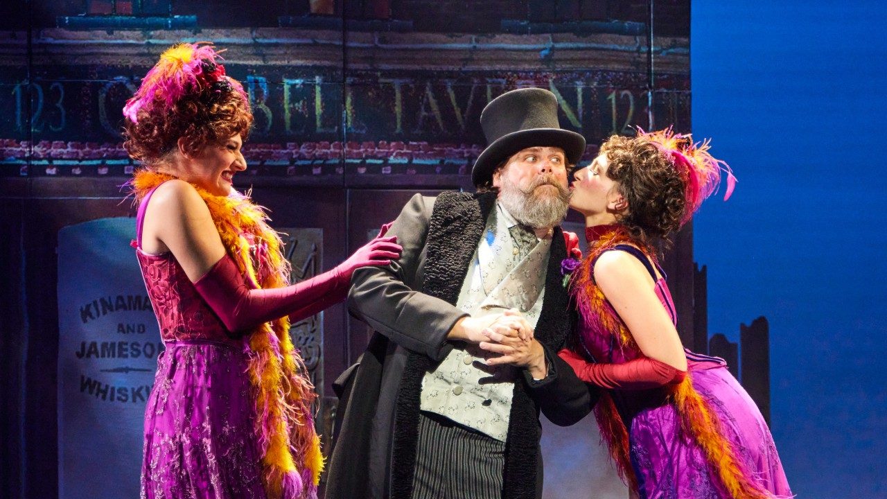  Sami Murphy, Michael Hegarty as Alfred P. Doolittle, and Ashley Agrusa in the national tour of "My Fair Lady." Photo by Jeremy Daniel. Two white women in early 1900s dresses of magenta and orange flank a middle aged white man wearing 1900s-inspired upper class clothing. The one on the right kisses him on the cheek.