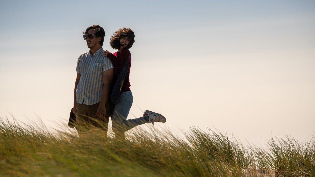  Vietnamese and Italian American singer and songwriter Julian Saporiti, at left, stands on a grassy beach dune at sunset, wearing brown ankle pants and a blue and white striped button-down short sleeved shirt. At right is his wife and collaborator, Emilia Halvorsen-Saporiti, a young Asian woman with shoulder length dark brown hair. She wears a red sweater and light blue skinny jeans, and holds onto his shoulders while kicking one foot up behind her and smiling towards the camera, he hair in her face from the wind.