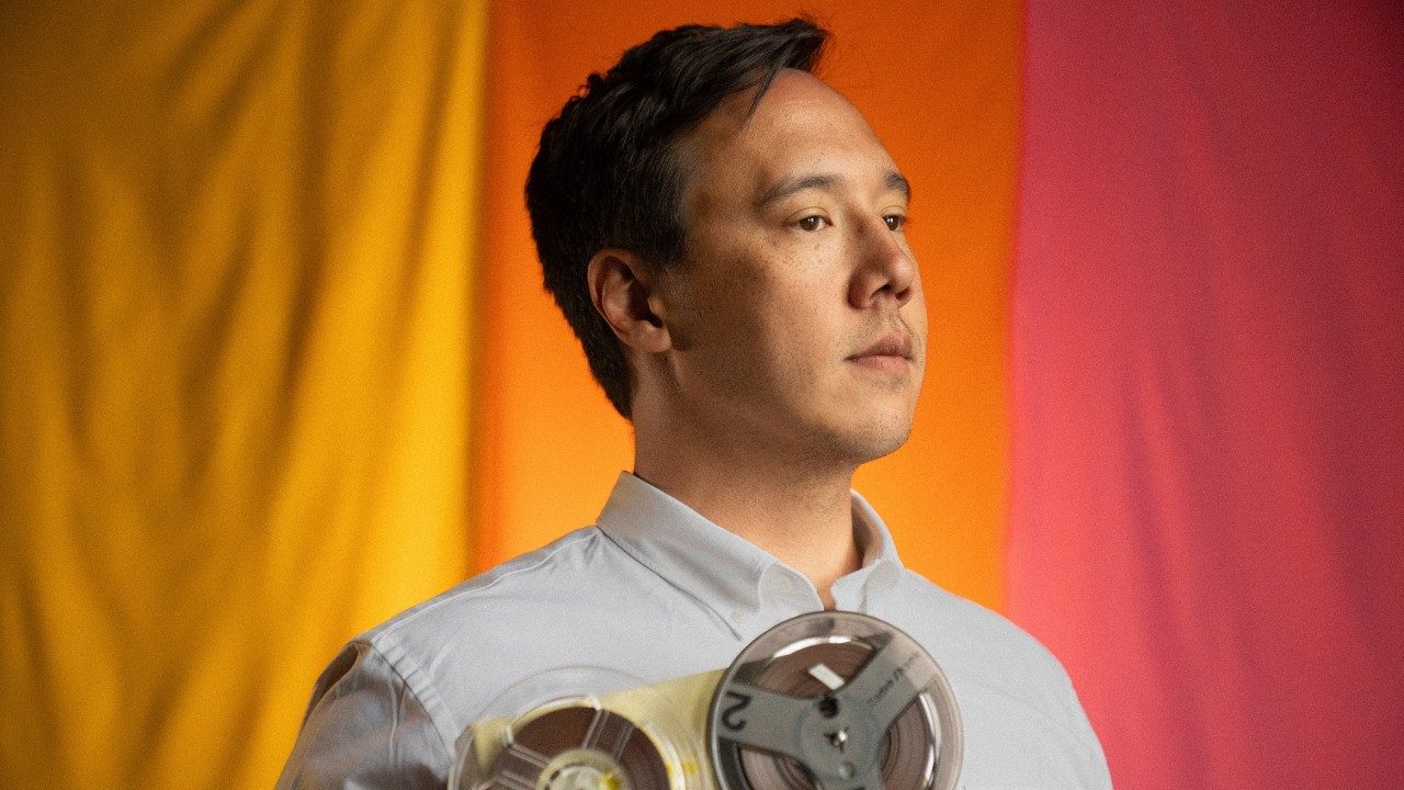 Vietnamese and Italian American singer and songwriter Julian Saporiti stands in front of a colorblocked pink, orange, and yellow background, and looks towards the right edge of the frame in a light grey button down shirt. He holds a vintage recording device.