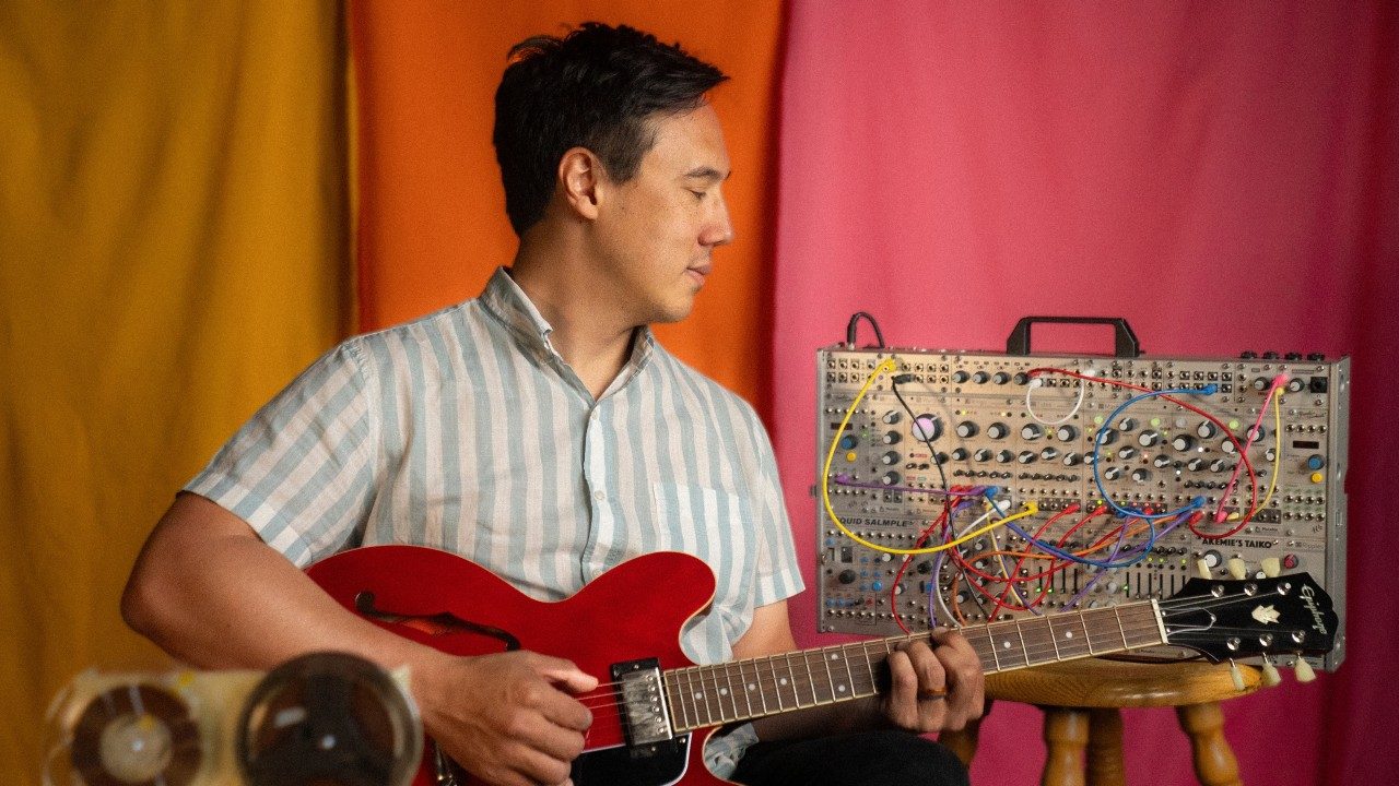  Vietnamese and Italian American singer and songwriter Julian Saporiti sits in front of a colorblocked pink, orange, and yellow background. He plays a red hollow body guitar in front of a mixing board and behind a vintage recording device, which is out of focus in the foreground. He wears a blue and white striped button down short sleeved shirt.