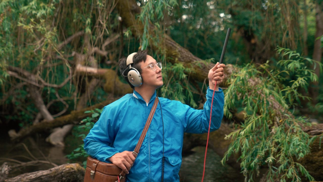  Vietnamese and Italian American singer and songwriter Julian Saporiti wears an electric blue windbreaker while he records birdsong and natural sounds in the field. He is in front of willow trees and vines, a stream behind him. He wears headphones and has a leather box slung over his shoulder, and holds a skinny rod microphone up, a red cord trailing down and into the leather box.