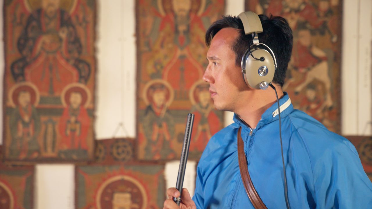  Vietnamese and Italian American singer and songwriter Julian Saporiti wears an electric blue windbreaker and beige over-ear headphones as he records. He speaks or sings into a skinny rod microphone in front of a white wall lined with what appears to be a series of small vintage religious art tapestries.