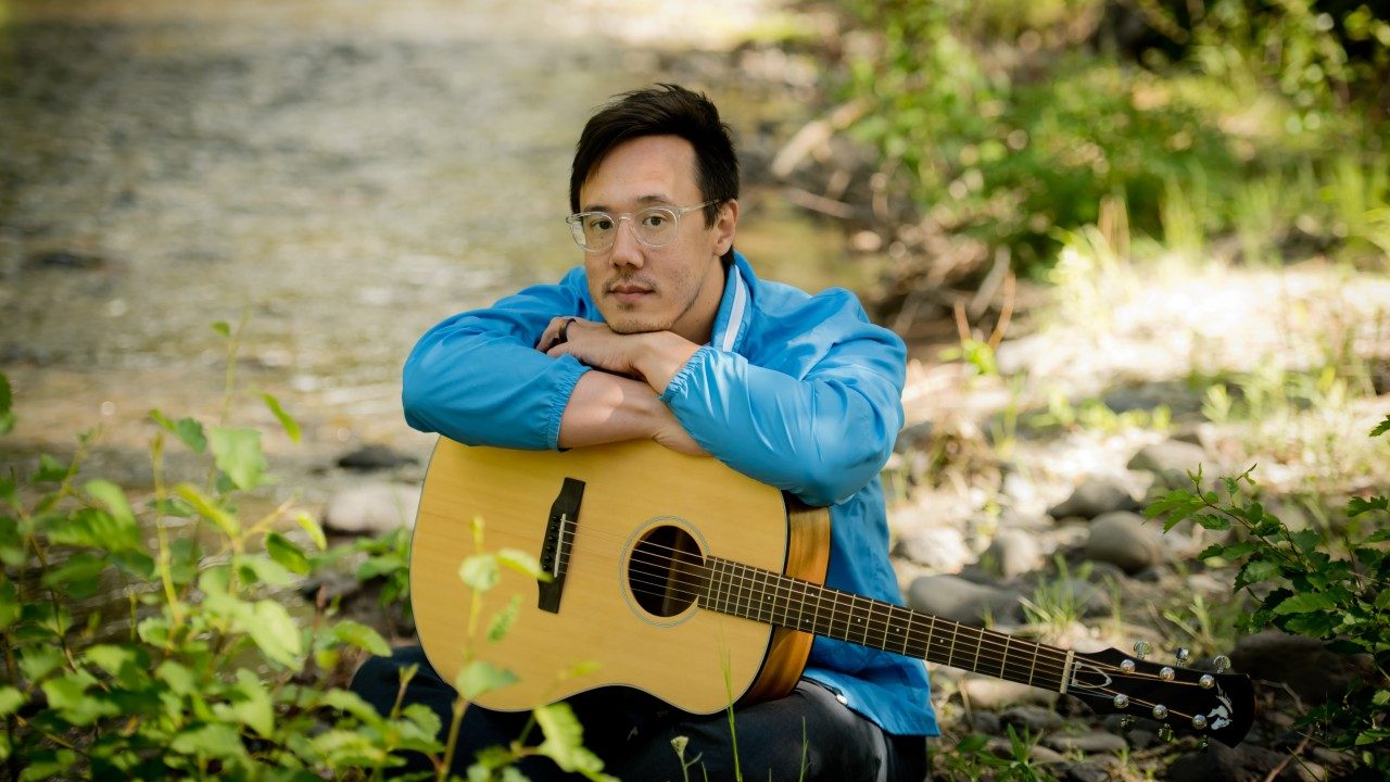  Vietnamese and Italian American singer and songwriter Julian Saporiti wears an electric blue windbreaker and clear rimmed glasses. He sits at the edge of a river or stream, an acoustic guitar in his lap, arms and chin resting on top. He gazes towards the camera.