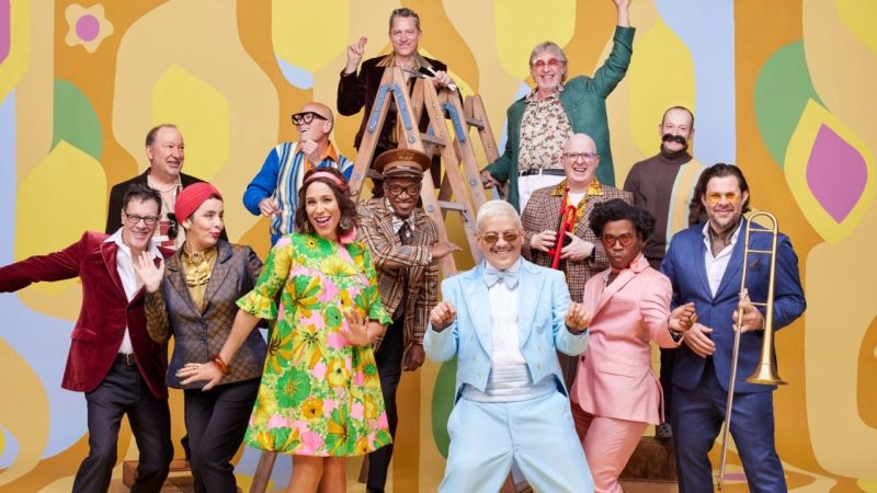  The members of Pink Martini wear '60s-inspired outfits and dance in front of a multicolored '60s-inspired backdrop, some of them holding their instruments.