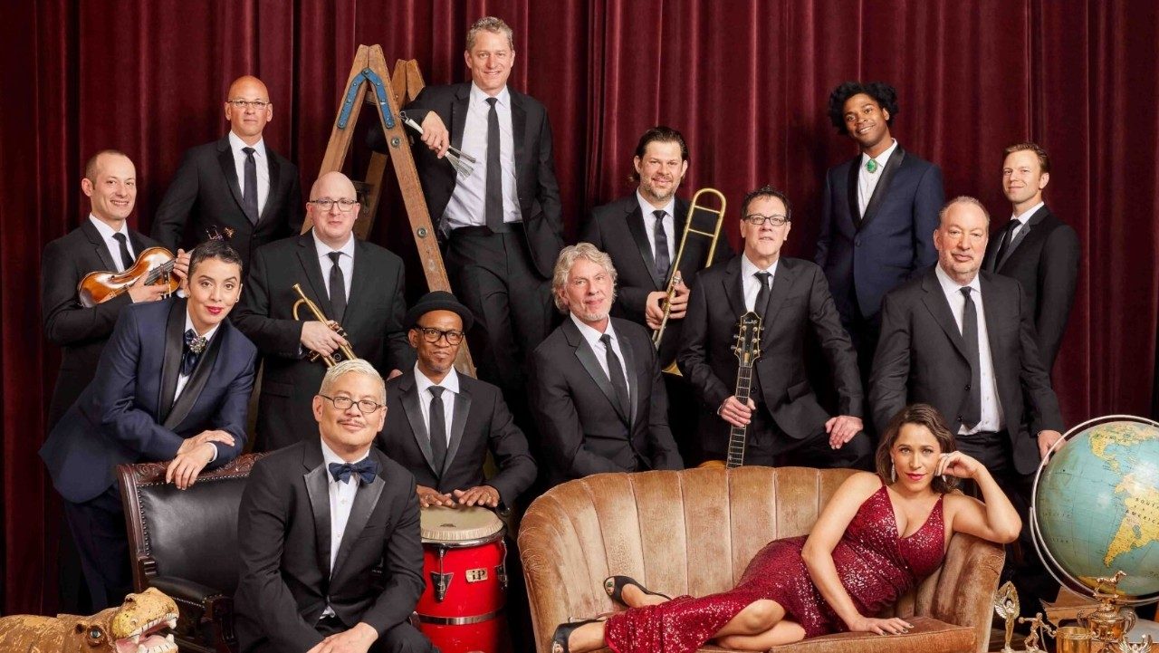  The members of Pink Martini wear black suites and gather in front of a red velvet curtain. In the foreground, singer China Forbes lounges on a blush pink settee in a long, red sequined dress Two members stand on a wooden ladder in the background, some members hold their instruments, and various props are visible, like a hippopotamos, globe, and various trophies. 