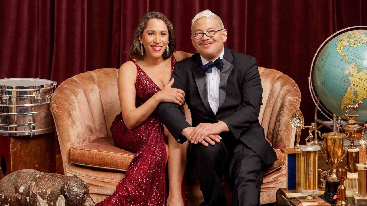  Singer China Forbes, at left, and pianist Thomas Lauderdale, at right, from the band Pink Martini sit on a blush settee in front of a red velvet curtain and smile towards the camera. Forbes is white woman with shoulder-length dark brown hair, and Lauderdale is an Asian man with bleached blonde short hair and glasses. She wears a red sequined floor length dress and he wears a black tux. Around them are various props, like a snare drum, a bear statue, a collection of trophies, and a globe.