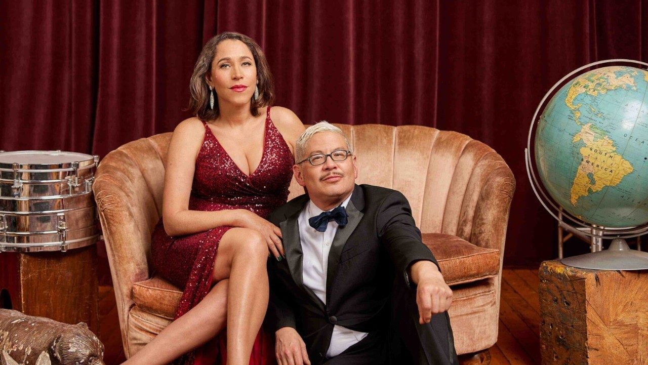  Singer China Forbes, at left, and pianist Thomas Lauderdale, at right, from the band Pink Martini sit on a blush settee in front of a red velvet curtain and look towards the camera. Forbes is white woman with shoulder-length dark brown hair, and Lauderdale is an Asian man with bleached blonde short hair and glasses. She wears a red sequined floor length dress and he wears a black tux. Around them are various props, like a snare drum, a bear statue, and a globe.