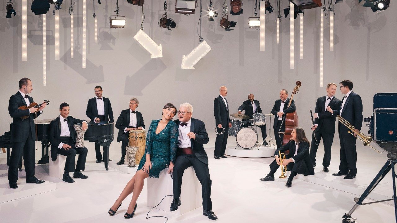  Members of Pink Martini wear tuxedos on a white stage set that looks similar to the Ed Sullivan Show set. In the background, the members stand or sit in a row with their instruments. In the foreground, singer China Forbes, a white woman with short dark brown hair, wears an emerald green knee-length dress and sits on a white cube with pianist Thomas Lauderdale, an Asian man with short bleached blonde hair wearing a tux and maroon cumberbund.