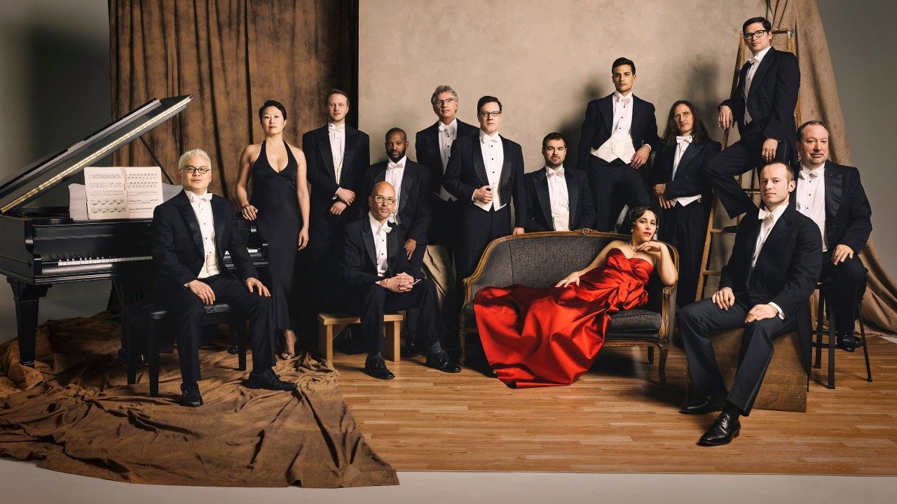  The members of Pink Martini wear black tuxedos or a black floor length gown and stand or sit on a photo set. In the middle of the group, singer China Forbes, a white woman with dark brown hair, lounges on a settee and wears a bright red gown. At far left, Thomas Lauderdale, an Asian man with short bleached blonde hair, sits at a grand piano.