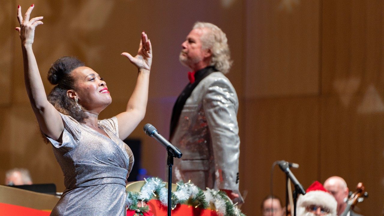  The Roanoke Symphony Orchestra performs its annual "Holiday Pops Spectacular" at the Moss Arts Center. The 2022 soloist, a Black woman in a silver dress, lifts both arms above her head and smiles towards the crowd. Behind her, conductor David Stewart Wiley, a white man with shoulder length greying blonde hair, looks over the orchestra in a silver blazer and red bow tie.