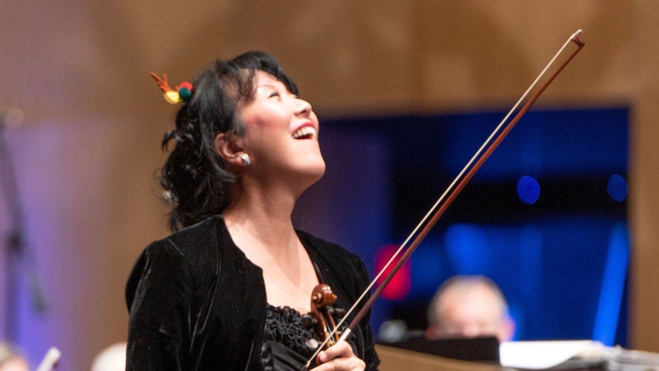  Roanoke Symphony Orchestra concertmaster Akemi Takayama, an Asian woman with dark brown hair pulled back into an updo, wears a black velvet jacket over a black dress and smiles into the house while holding her violin and bow.