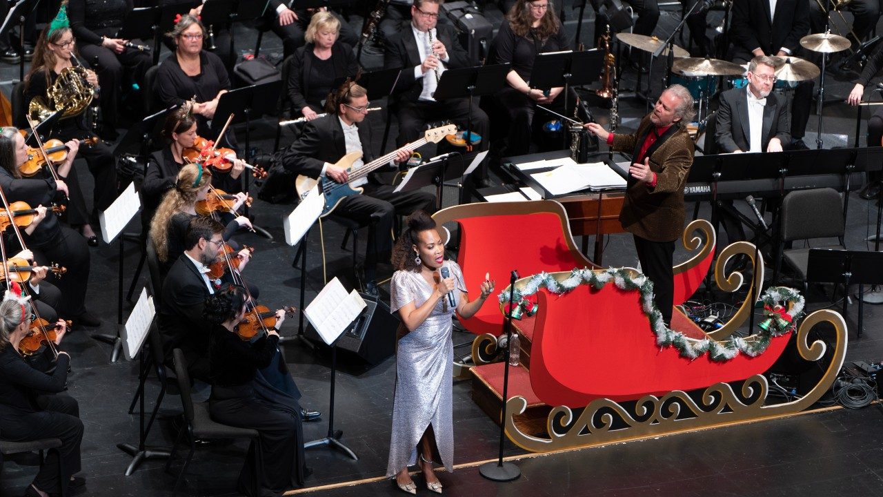  The Roanoke Symphony Orchestra performs its annual "Holiday Pops Spectacular" at the Moss Arts Center. In the foreground, the 2022 soloist, a Black woman in a floor-length silver dress, sings while David Stewart Wiley conducts from a red sleigh nearby.