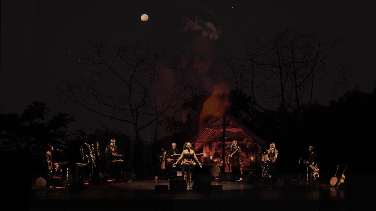 Cast members of "Small Island, Big Song" sing, dance, and play instruments on stage in front of a large projection on a screen. The projection shows a yurt with a campfire in front, leafless trees, stars, and the moon visible in the sky above it. Two women's faces are transposed on top of the landscape scene.