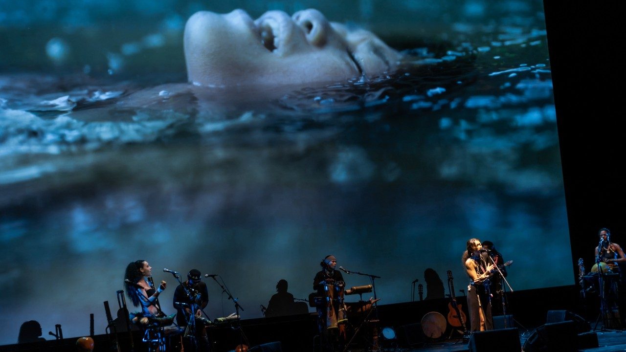 Performers from "Small Island, Big Song" sing and play instruments on stage. Behind them is a large projected image of an Indigenous person floating in water in the rain.