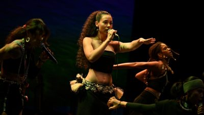 A young brown woman with long wavy hair pulled back into a ponytail sings on stage while other women dance nearby. She wears a traditional belt with large fringed tassels around her hips.