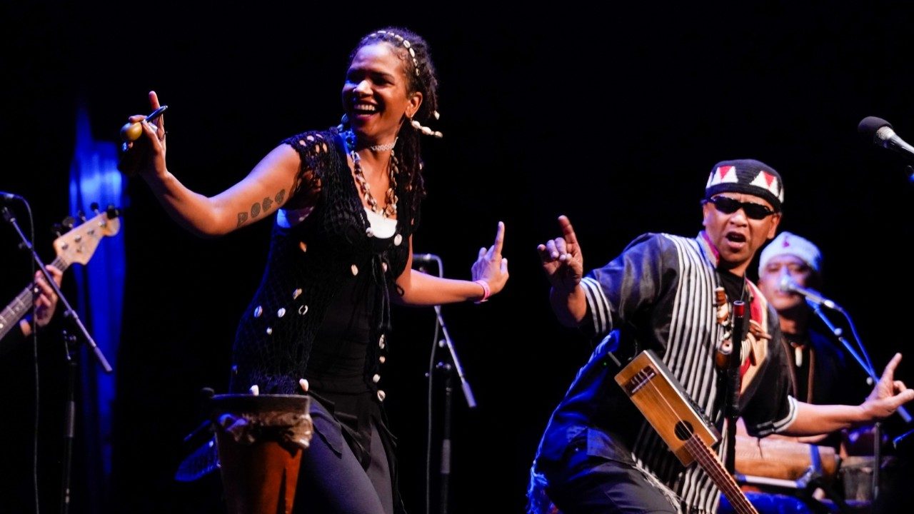  Cast members from "Small Island, Big Song" wear traditional costumes and accessories, and dance on stage. At left is a light-skinned Black woman, and at right is a brown man in a traditional hat and sunglasses with a traditional instrument, like a ukulele but with a rectangular body, hanging from his shoulder by its strap.