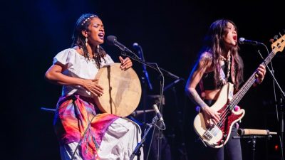 Two young AAPI women wearing traditional dress and accessories perform during "Small Island, Big Song." At left, a Black woman with a white dress, pink and orange scarf tied around her waist, and puka shell jewelry sings and plays a large hand drum. At right, a woman with long brown hair wears a black crop top, black pants, and traditional jewelry and plays a red and white bass guitar.
