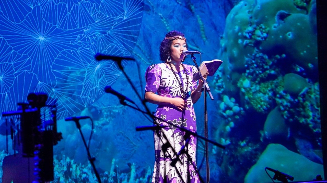  A young AAPI woman wears a long, purple, patterned dress and traditional jewelry and reads poetry. Behind her is a large projection of coral and a ginko leaf pattern.