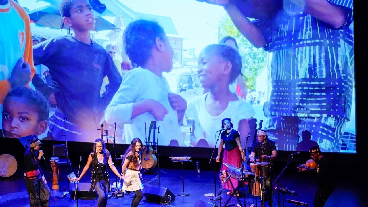  Cast members from "Small Island, Big Song" sing, dance, and play instruments on stage in front of a large projection of children from Pacific and Indian Ocean islands.