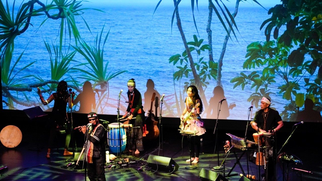  Cast members from "Small Island, Big Song" sing and play instruments in front of a large projection of a view of the ocean through green vegetation on a Pacific or Indian Ocean island.