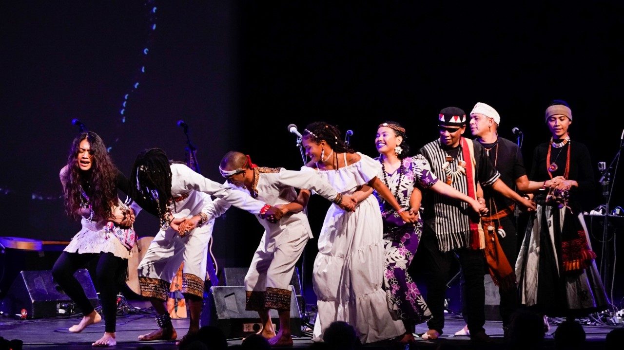  Cast members from "Small Island, Big Song" join hands and sing and dance in a line, all wearing traditional costumes from their native countries.