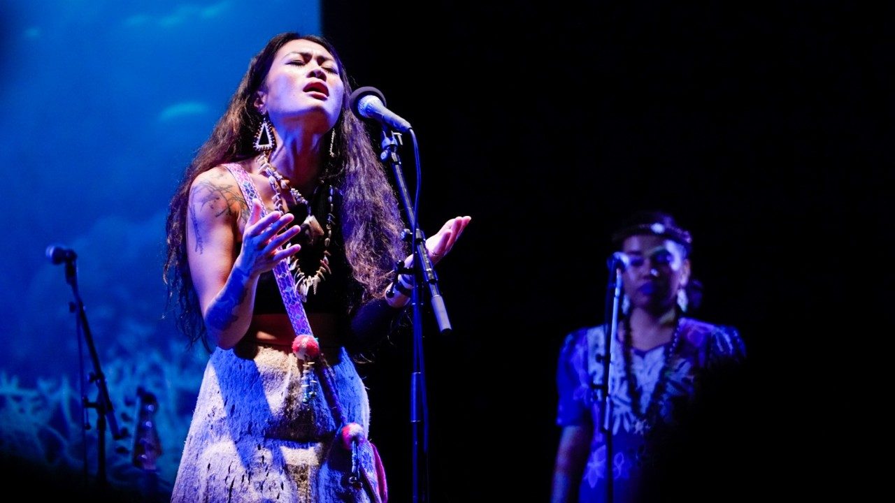  A young woman with long brown hair sings into a microphone on stage during a performance of "Small Island, Big Song." She wears a traditional costume, her hands and face lifted gently upwards. In the background, another woman in traditional dress stands at a microphone.