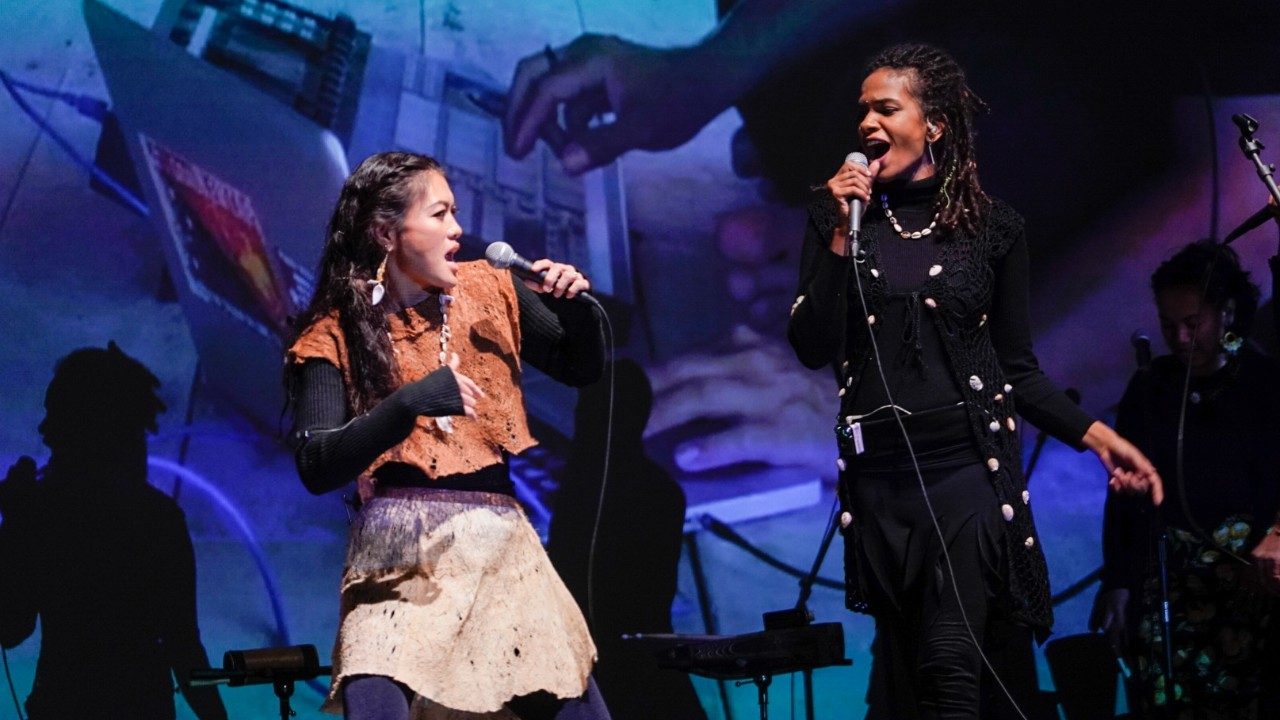  Two young AAPI women wearing traditional costumes and accessories sing and dance on stage during a performance of "Small Island, Big Song." Behind them is large projected imagery from Pacific and Indian Ocean islands.