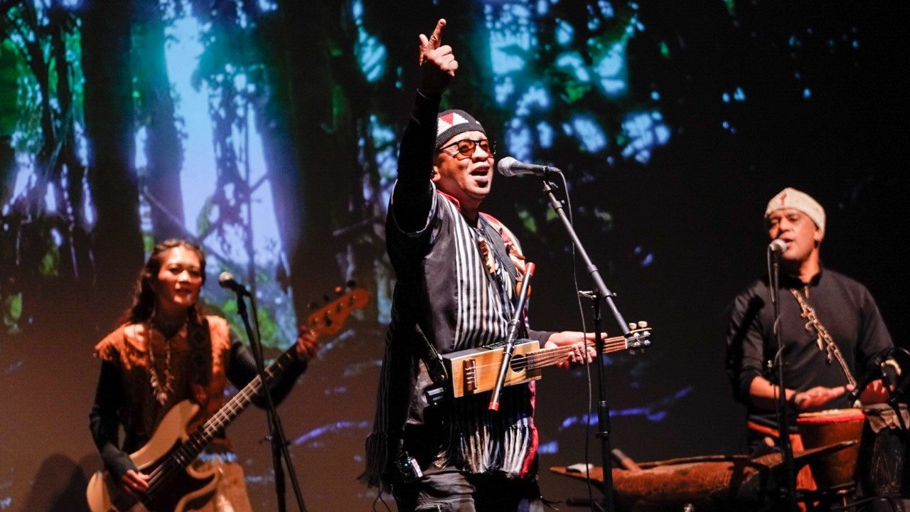   Three singers and musicians from "Small Island, Big Song" perform on stage. In the middle foreground is a middle aged brown man wearing traditional dress and hat. He holds an instrument that resembles a ukulele but with a thin rectangular body and points one finger up in the air.