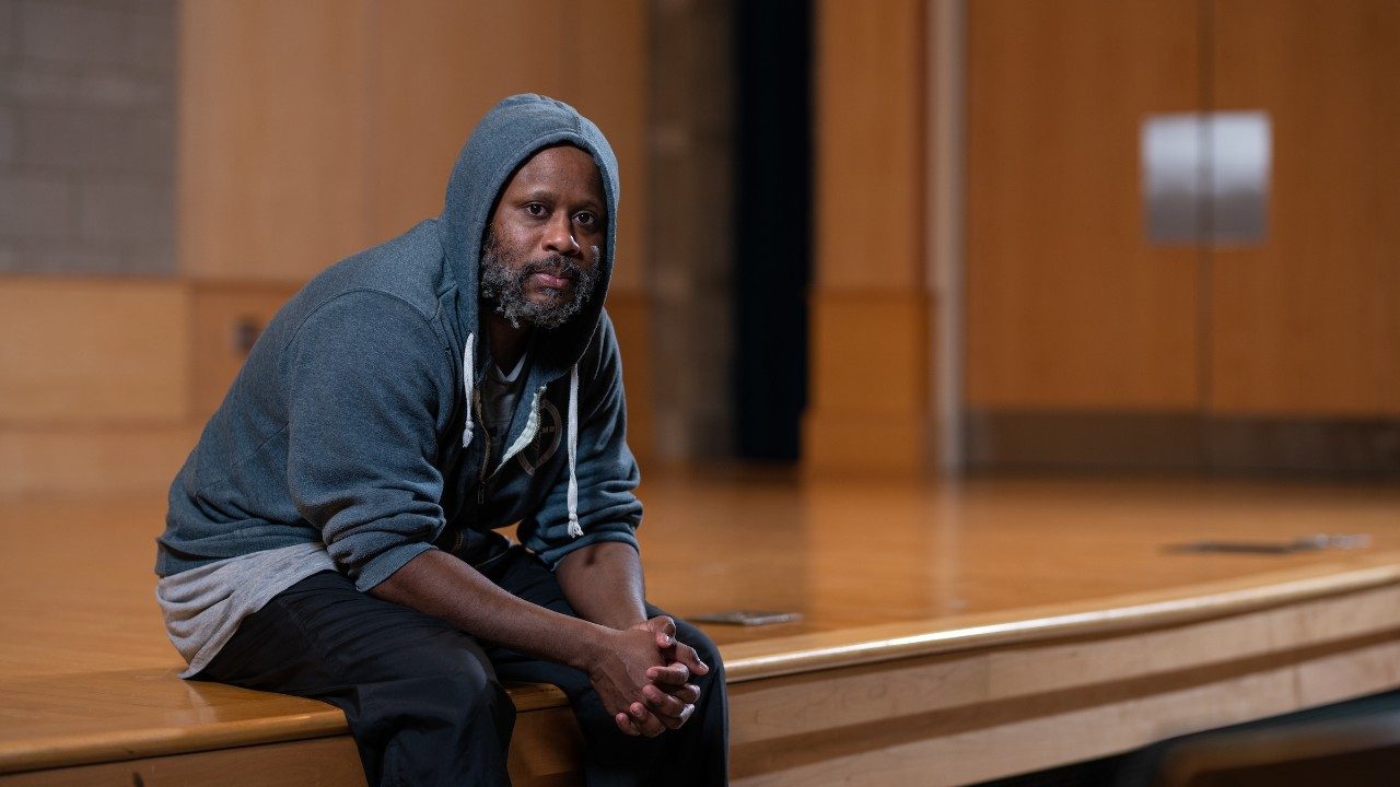  Beat boxer and voice percussionist Shodekeh, a Black man with a salt and pepper beard wearing a grey hoodie with the sleeves pushed up and hood on his head, sits on the edge of a stage and looks towards the camera.