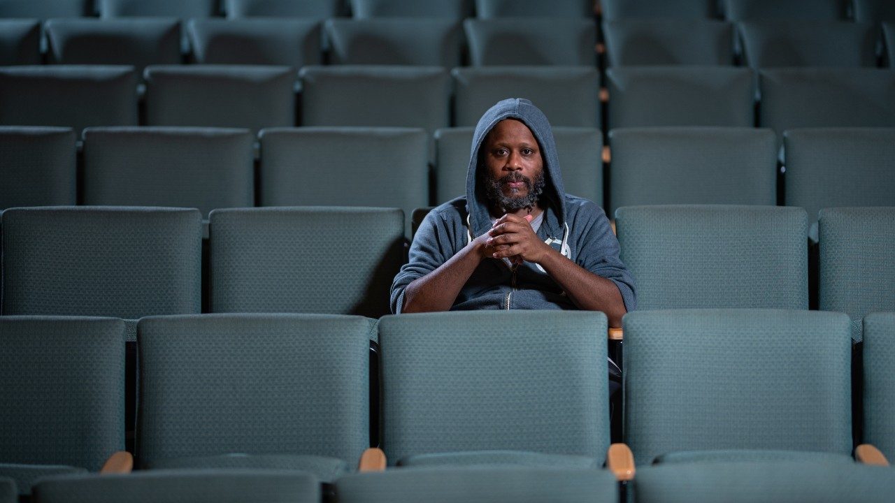  Beat boxer and voice artist Shodekeh, a Black man with a salt a pepper beard, wears a grey hoodie with the sleeves pushed up and hood over his head. He sits in the sage green seats of an empty auditorium and looks towards the camera.