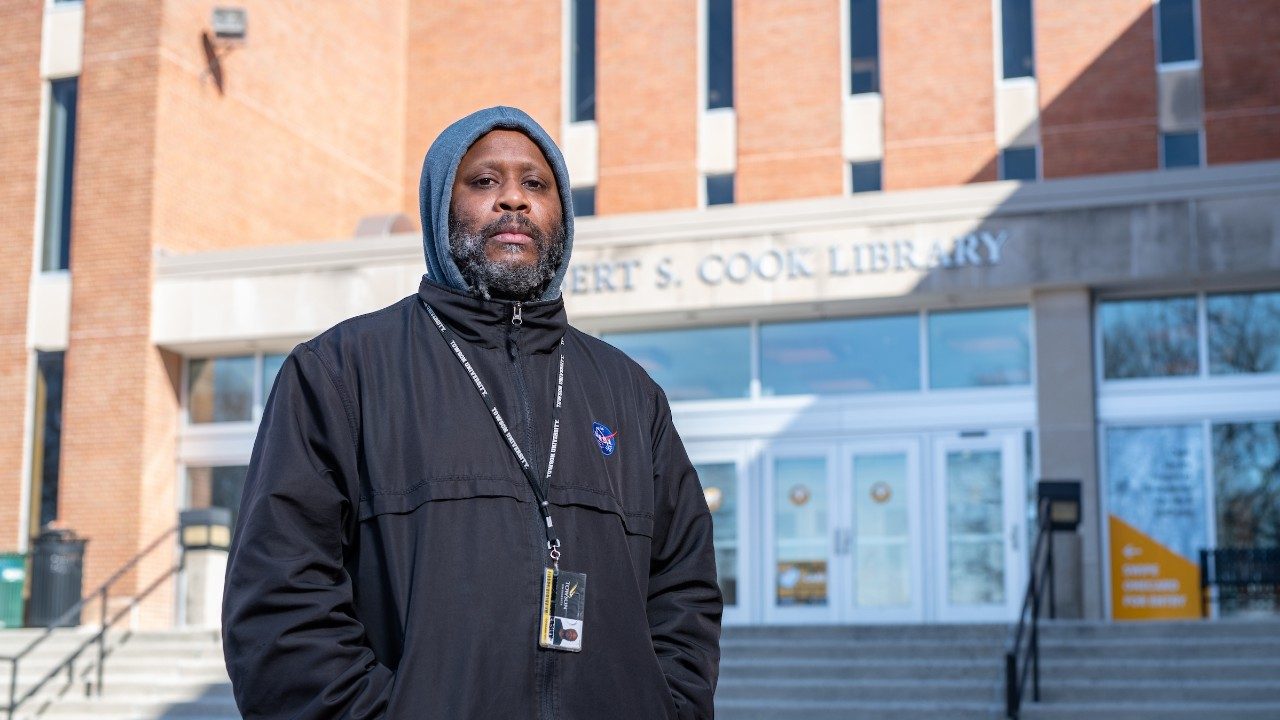  Beat boxer and voice artist Shodekeh, a Black man with a salt a pepper beard, wears a grey hoodie with the hood over his head and a black jacket. He stands in front of the Robert S. Cook Library stairs at Towson University with his hands in his pockets, the sun pouring into the frame at a diagonal angle.