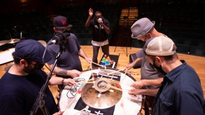  The members of Sō Percussion, four white men wearing casual clothes, hats, and face masks, perform on the same drum and cymbal contraption, while, nearby, hip-hop and breath artist Shodekeh, a Black man wearing a black hoodie and a light blue face mask, performs into a microphone.