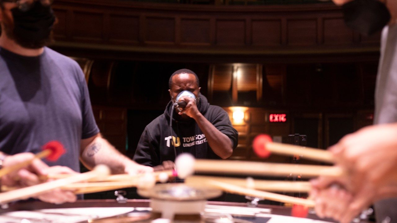  Members of Sō Percussion perform out of focus in the foreground, while beatboxer and breath artist Shodekeh, a Black man in a black hoodie with a light blue face mask, performs nearby.