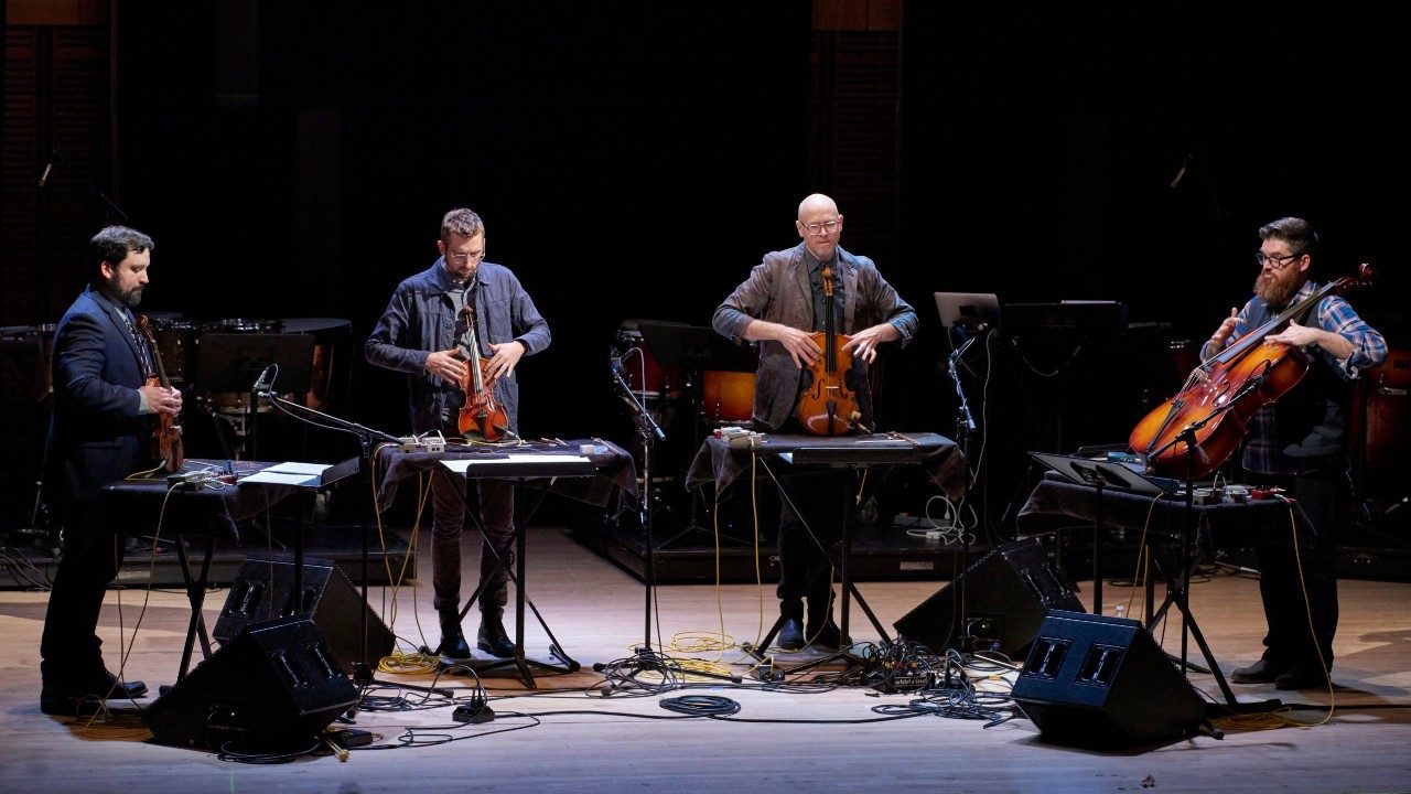  Members of Sō Percussion, four white men, perform on stage using the instruments of a string quartet for their percussion.