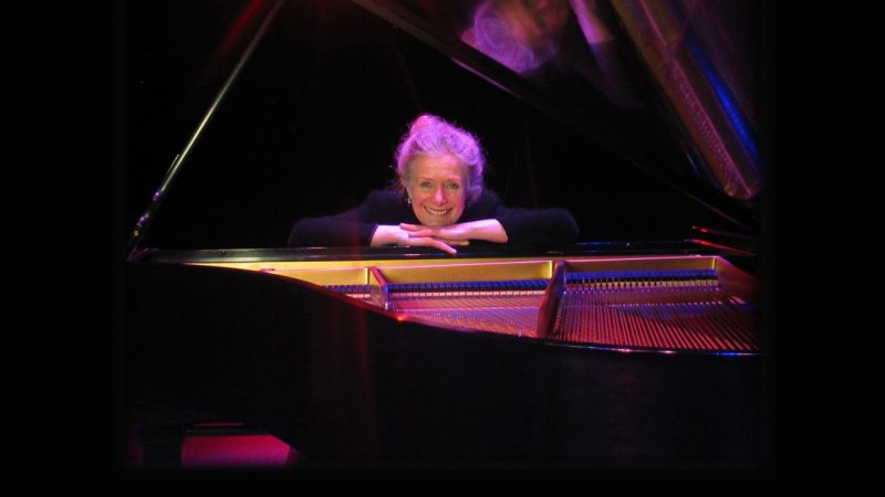 Pianist Barbara Nissman, a white woman with blonde hair pulled into a bun, leans onto an on open grand piano, her chin rested on her folded hands.