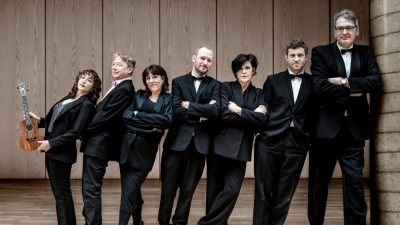  Members of the Ukulele Orchestra of Great Britain, three white women and four white men, stand in a line wearing all black. The women are order from shortest to tallest, left to right, and the men are ordered the same way., and they all lean right towards the tallest man. The woman on the far left holds a ukulele.