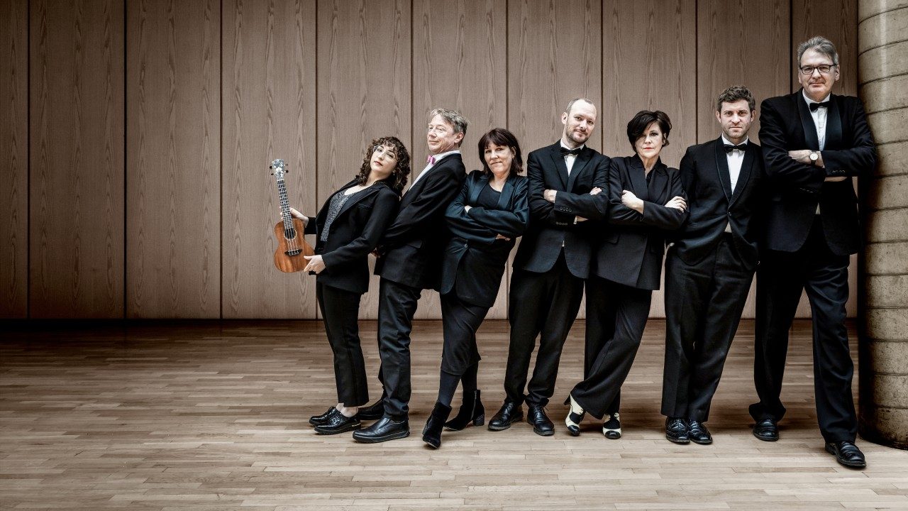  Members of the Ukulele Orchestra of Great Britain, three white women and four white men, stand in a line wearing all black. The women are order from shortest to tallest, left to right, and the men are ordered the same way., and they all lean right towards the tallest man. The woman on the far left holds a ukulele.