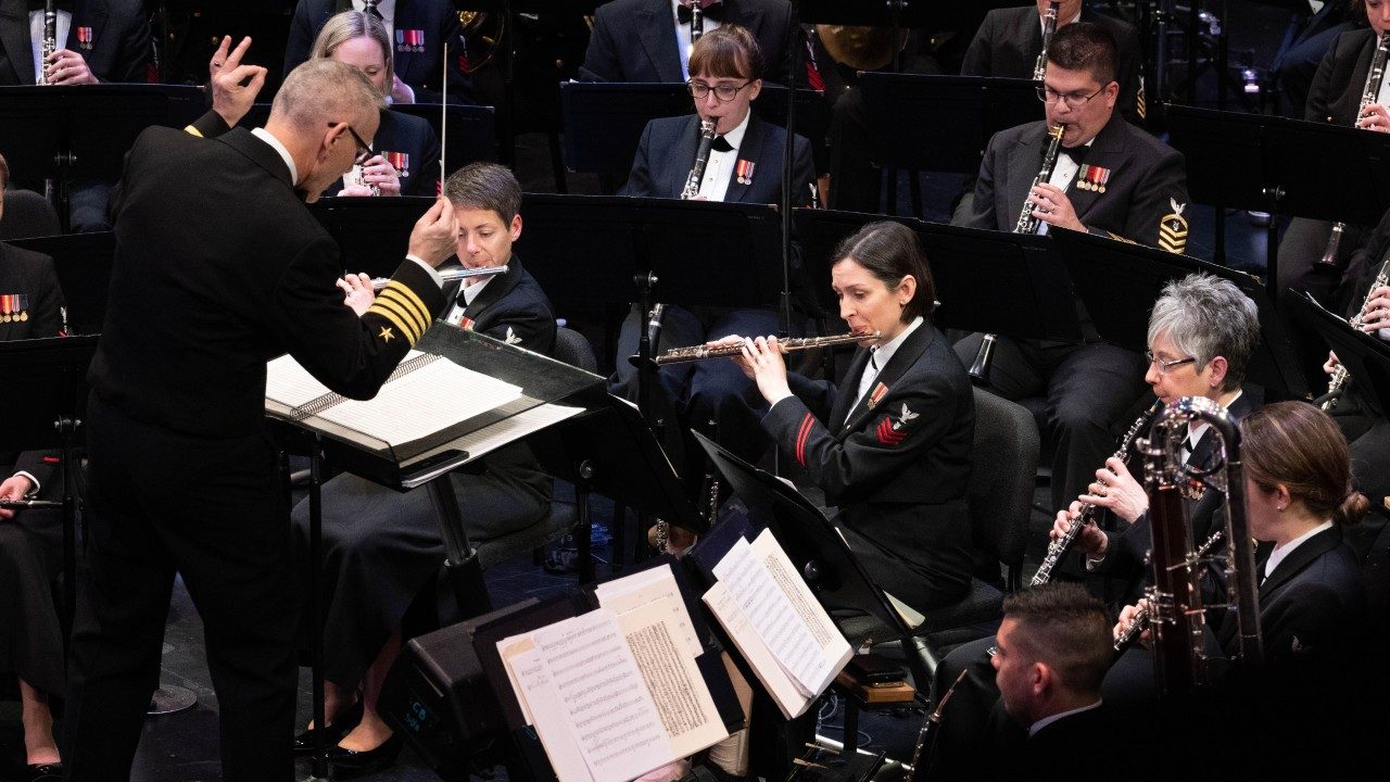 The United States Navy Band performs, its members wearing black uniforms and playing wind instruments.