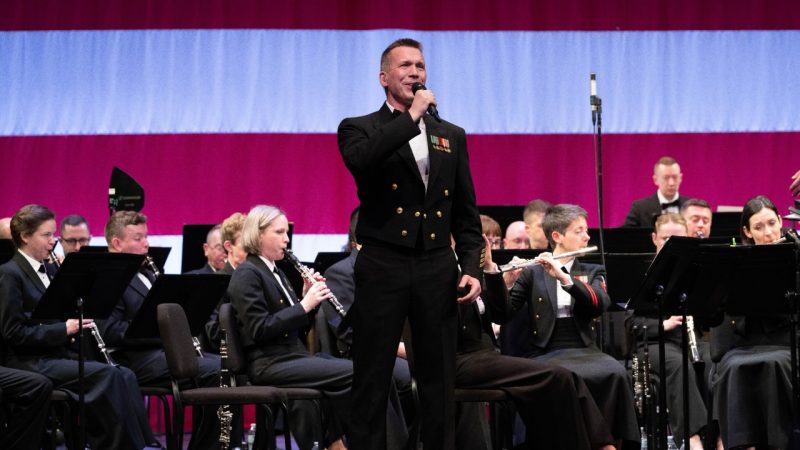 Chief Musician Bill Edwards, from Bowie, Md., sings with the U.S. Navy Concert Band at Pittsburg State University during the band’s 2023 national tour. He is a white middle aged man with a high and tight cut wearing a black uniform and he sings into a microphone, a large US flag on the wall behind the band.