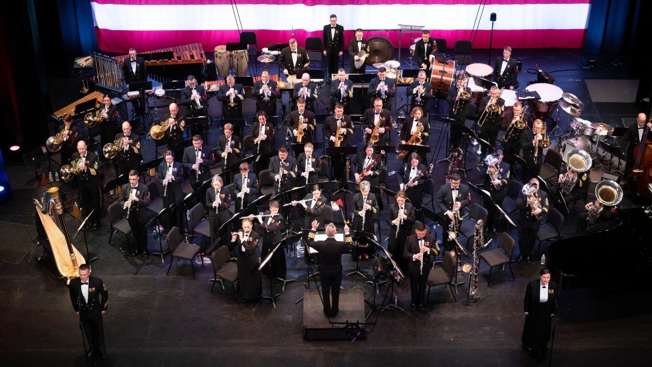 The U.S. Navy Concert Band performs at Pittsburg State University during the band’s 2023 national tour. Band members wear navy blue uniforms and play wind instruments. The photo was taken from above looking down on the stage.