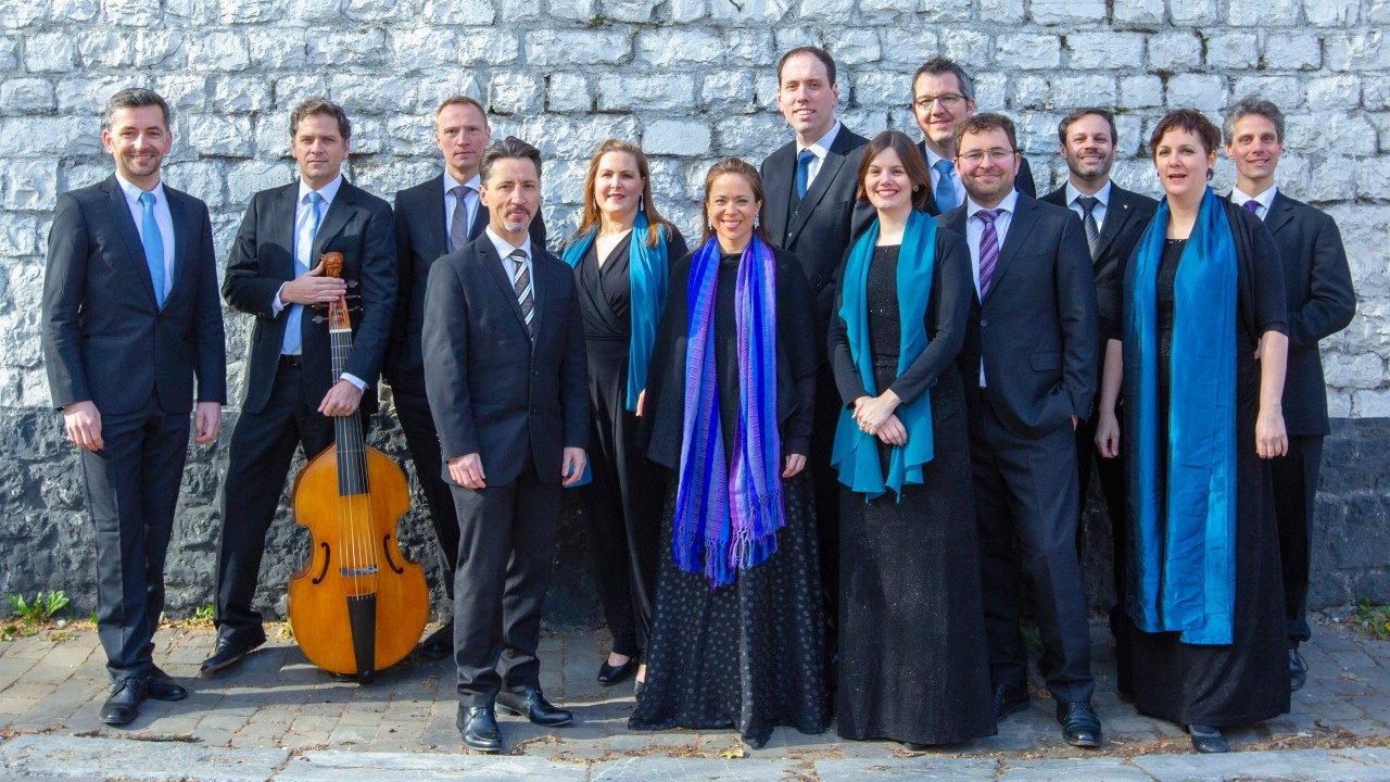  Members of Vox Luminis wear all black and stand in a row. The women in the group wear teal and purple scarves draped around their necks, and one man holds a cello-like instrument.