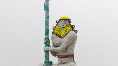 A statue of a male figure with a gold helmet grips a green pole. Michael Rakowitz; "The invisible enemy should not exist – Seated Nude Male Figure, Wearing Belt Around Waist (IM77823) (Recovered, Missing, Stolen Series)," 2018 (detail); Middle Eastern packaging and newspapers, glue, and cardboard; 37.8 x 28.7 x 28.7 inches; courtesy of the artist and Barbara Wien Gallery